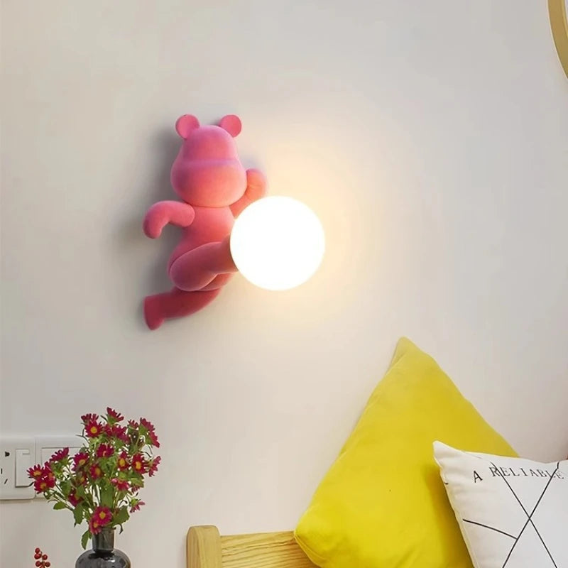 Różowa lampa miś SANDYHA z kulistym światłem zamontowana na ścianie nad łóżkiem z żółtą poduszką