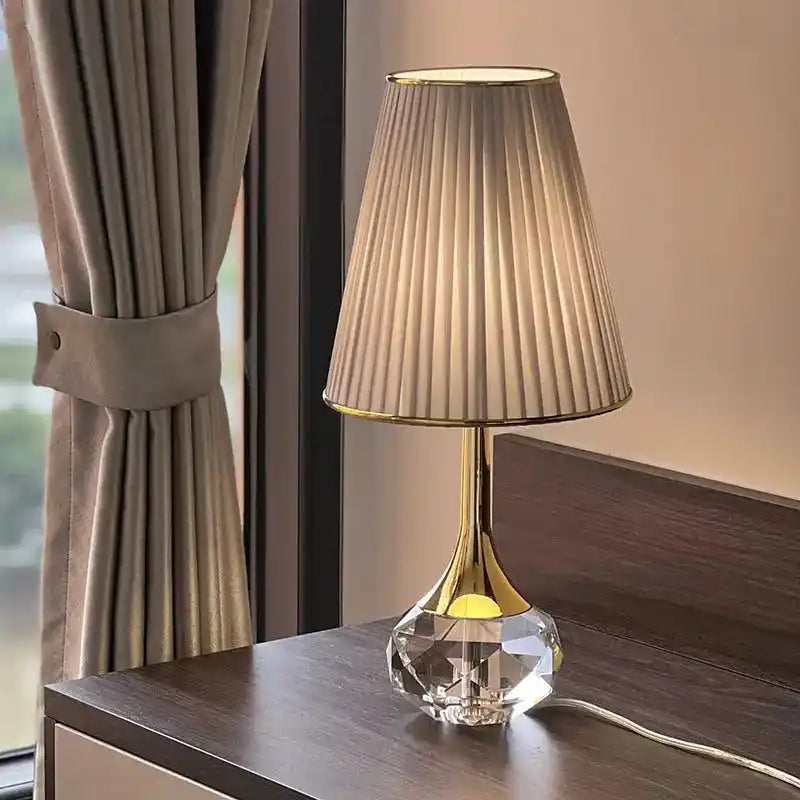 Lampa stołowa Moss z kryształową podstawą i plisowanym abażurem na ciemnym stoliku nocnym