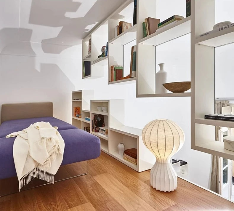 Designerska lampa Barcato na drewnianej podłodze obok fioletowej sofy i białych półek z książkami