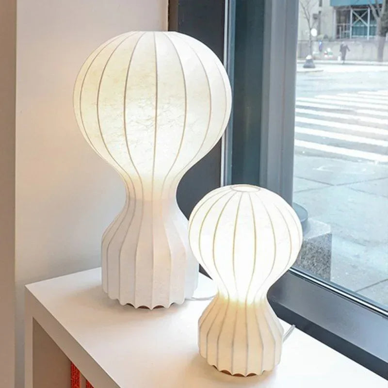 Designerska lampa Barcato w dwóch rozmiarach o oryginalnym kształcie stojąca na stoliku przy oknie