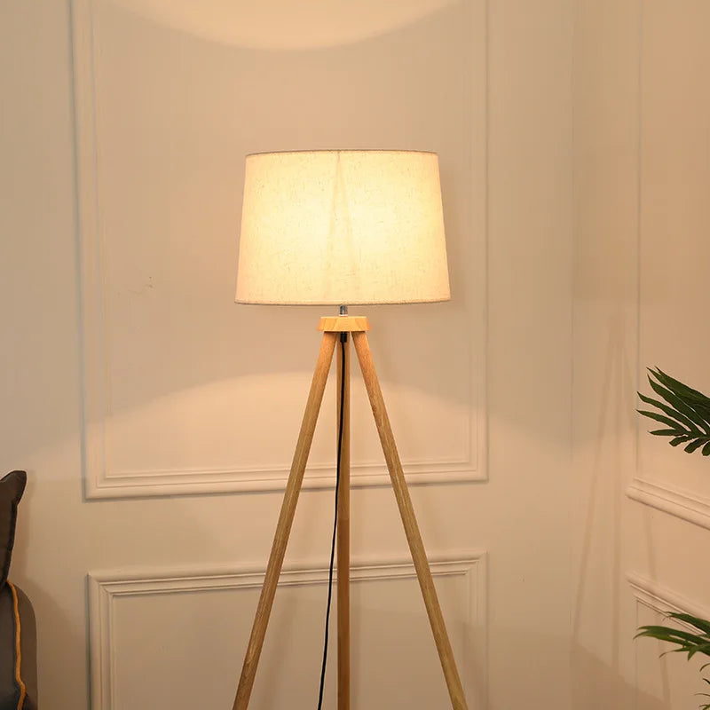 Elegancka lampa stojąca do salonu z jasnym abażurem i drewnianą podstawą na trójnogu
