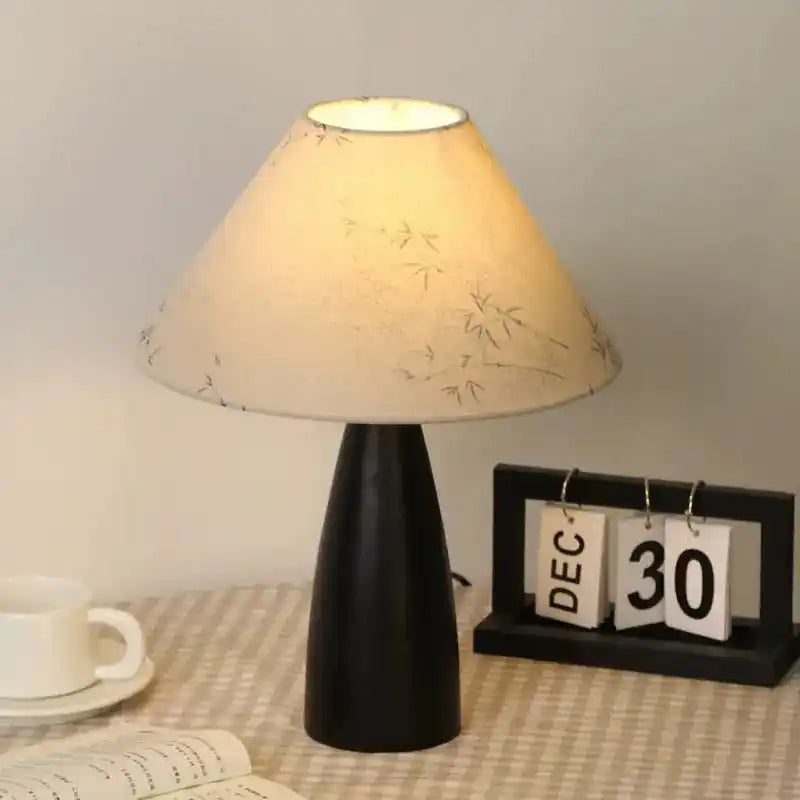 Lampa stołowa z jasnym abażurem i czarną podstawą ustawiona na stoliku z kalendarzem i filiżanką