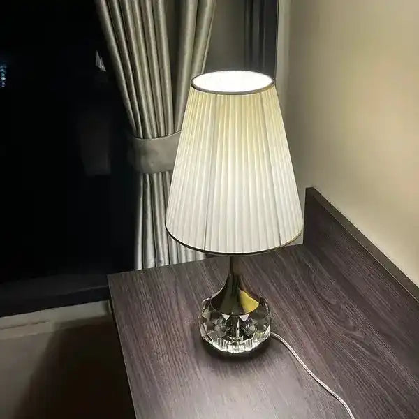 Elegancka lampa stołowa Moss z kryształową podstawą i białym plisowanym abażurem na ciemnym stole