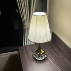 Elegancka lampa stołowa Moss z kryształową podstawą i białym plisowanym abażurem na ciemnym stole