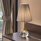 Lampa stołowa Moss z kryształową podstawą i plisowanym abażurem na ciemnym stoliku nocnym