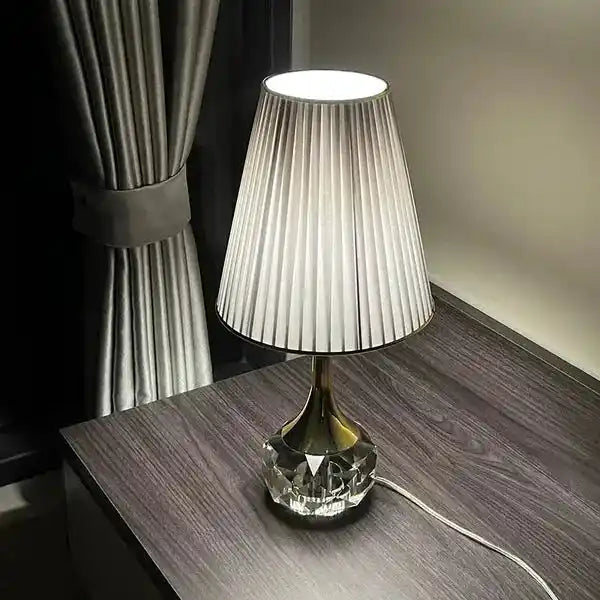 Lampa stołowa Moss z plisowanym białym abażurem i szklaną podstawą na ciemnym drewnianym stole