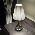 Lampa stołowa Moss z plisowanym białym abażurem i szklaną podstawą na ciemnym drewnianym stole