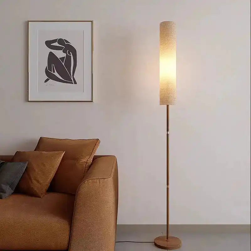 Stylowa lampa do wnętrz stojąca z jasnym abażurem obok brązowej kanapy i plakatu na ścianie